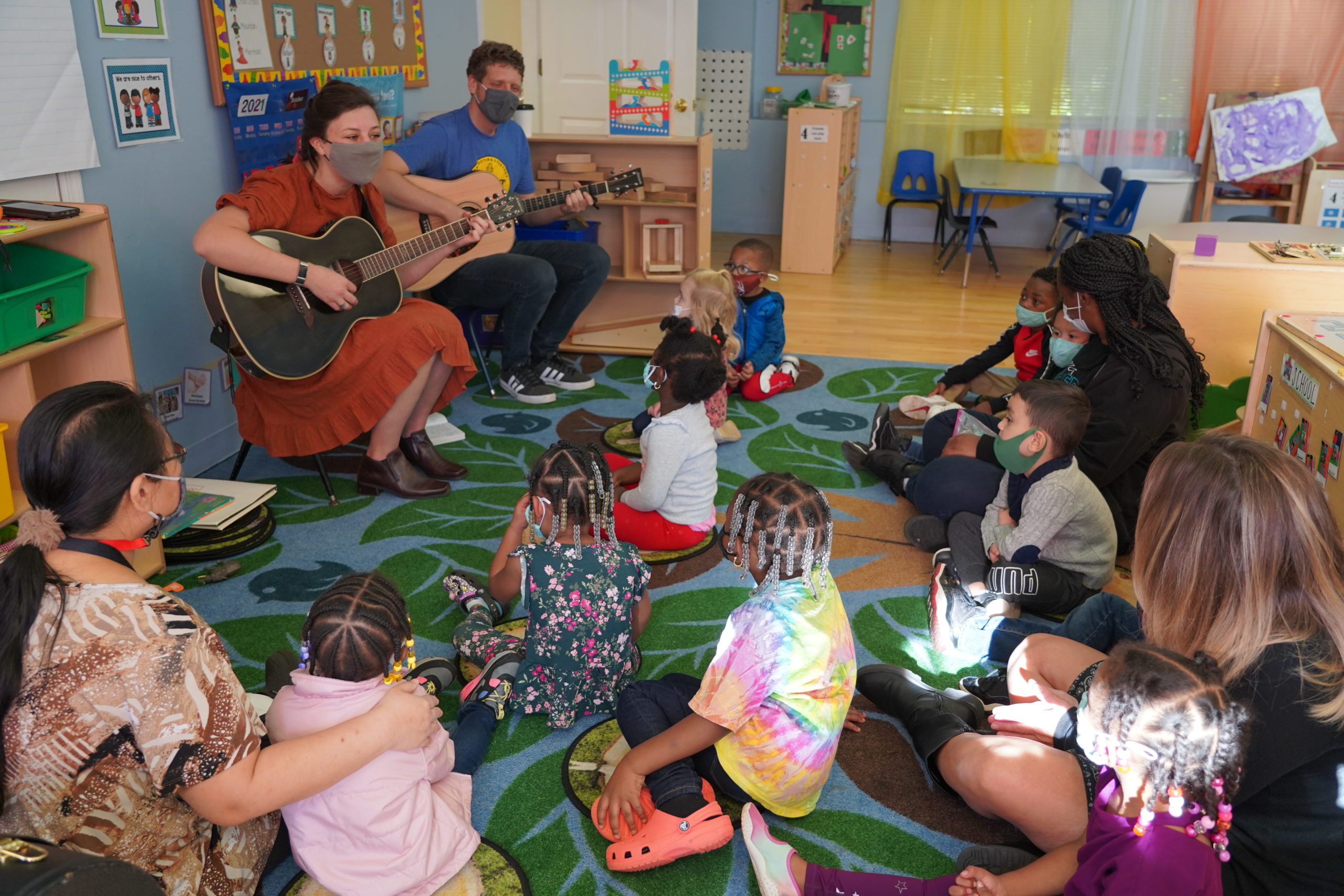 Two music teachers playing guitar in front of a group of young children sitting on a carpet. 